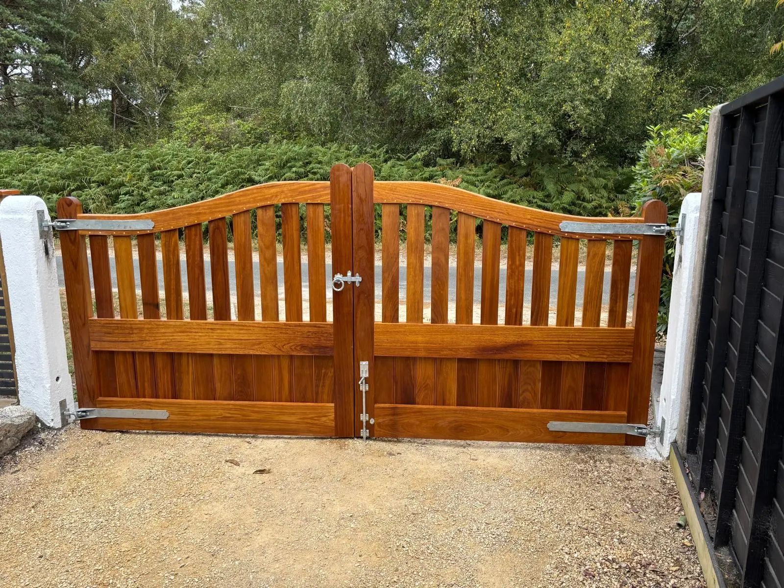 Swept-top hardwood driveway gates with vertical bar tops and rich oiled finish before woodland